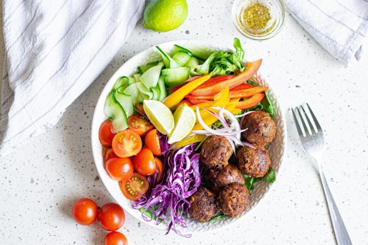 Lentil meatballs served in a bowl with cabbage, peppers, tomatoes, cucumber, red onion, and limes.