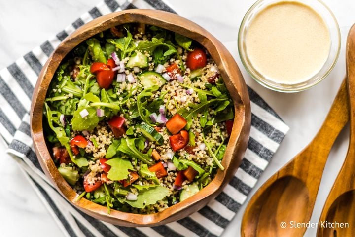 Mediterranean quinoa bowl with quinoa, arugula, red peppers, tomatoes, and hummus dressing in a wooden bowl.