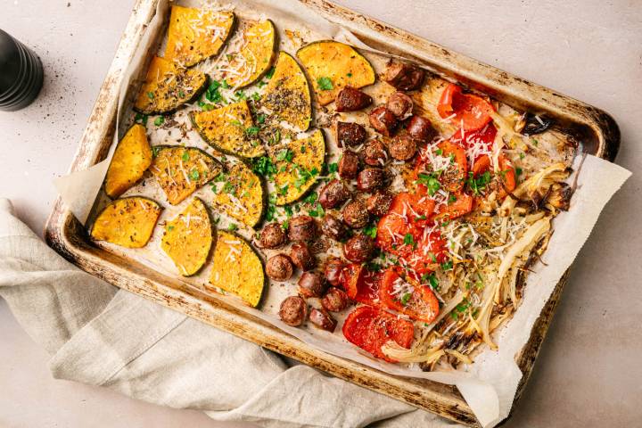 Roasted vegetables on a baking tray with savory sausage. Includes sliced pumpkin, tomatoes, and onions, garnished with herbs and grated cheese. Warm, rustic feel.