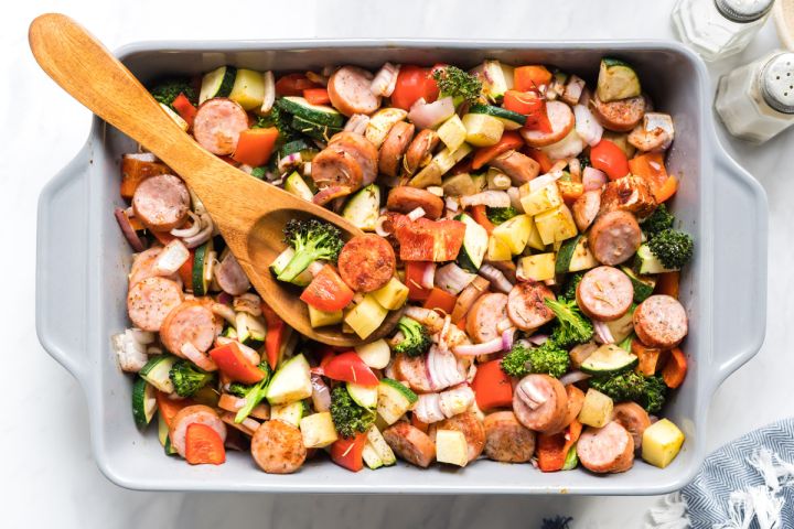 One pan sausage, potato, and veggie in a baking dish with a wooden spoon.
