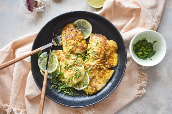 Pan fried garlic lime tilapia on a plate with fresh limes, green onions, and garlic.