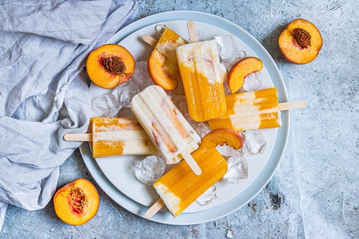 Peaches and cream popsicles with fresh peaches, honey, and Greek yogurt on a plate with ice and fresh peaches.