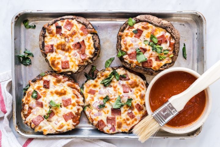Portobello mushroom pizzas on a baking sheet with marinara sauce, melted cheese, basil, ham, and onions.