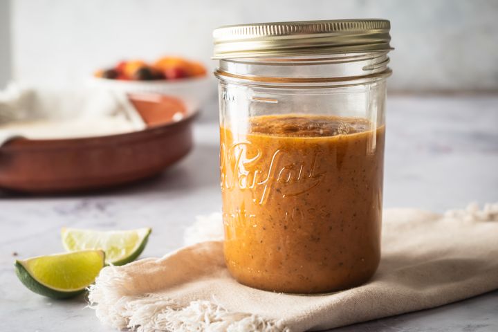 Homemade ranchero sauce in a glass jar with limes and tomatoes on the side.