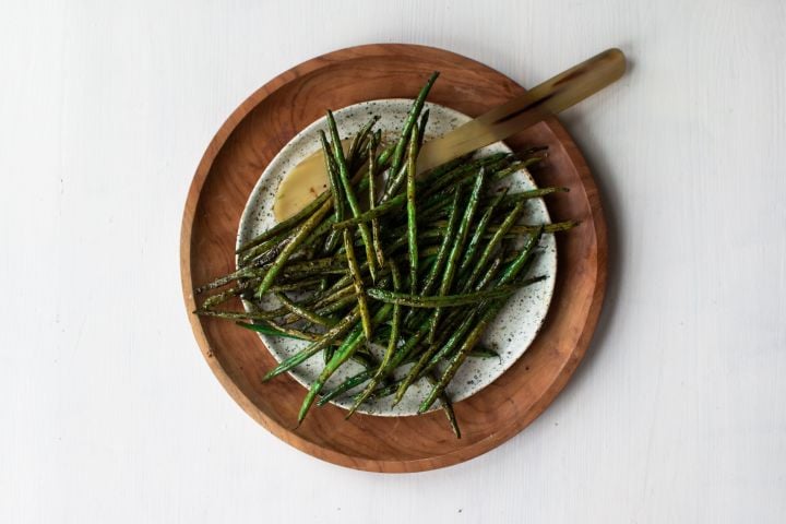 Roasted green beans with olive oil, balsamic vinegar, garlic powder, salt, pepper, and Italian seasoning on a wooden plate with a spoon. 