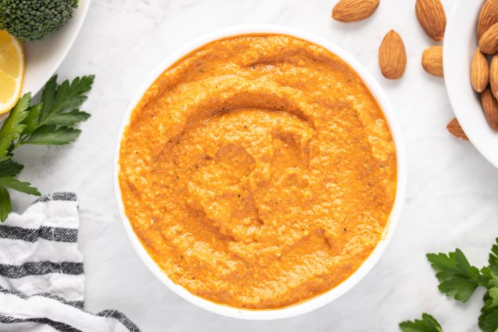 Romesco sauce with blended almonds, tomatoes, and red peppers in a small white bowl with parsley and lemon on the side.