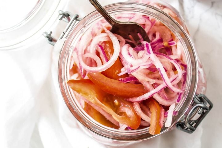Salsa criolla with red onions, tomatoes, and lime juice in a glass jar with a spoon.