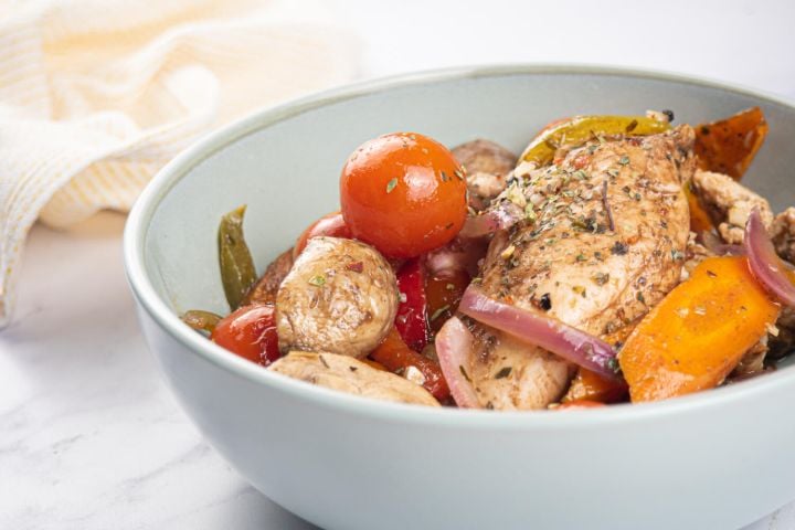Sheet pan balsamic chicken and vegetables in a white bowl with tomatoes, mushrooms, carrots, garlic, and spices.