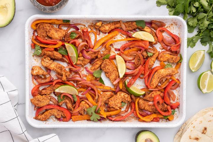 Sheet pan chicken fajitas with chicken breast, peppers, onions, and limes on a baking sheet .
