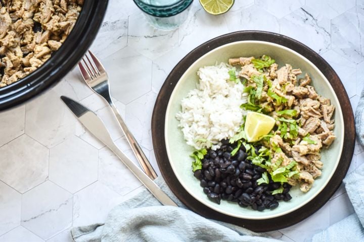 Slow cooker garlic cuban pork shredded on a plate with black beans, white rice, lime, and cilantro.