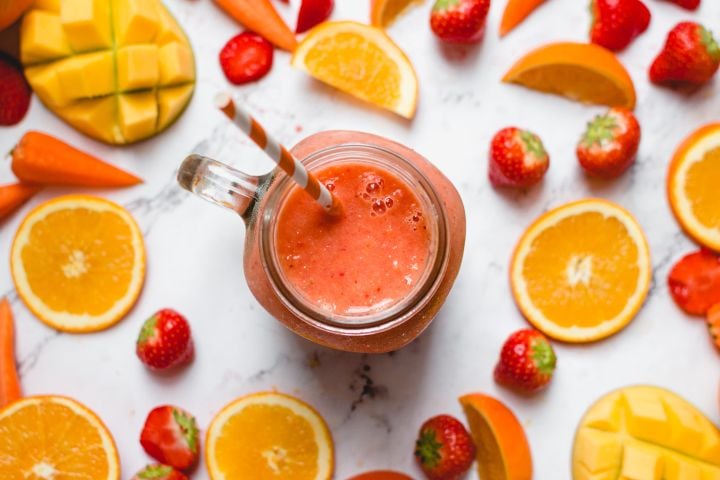 Strawberry mango smoothie in a glass with fresh strawberries, mangoes, and carrots on the side.
