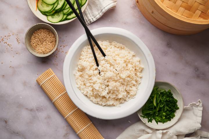 Sushi rice in a bowl with a bamboo mat and sliced vegetables on the side.