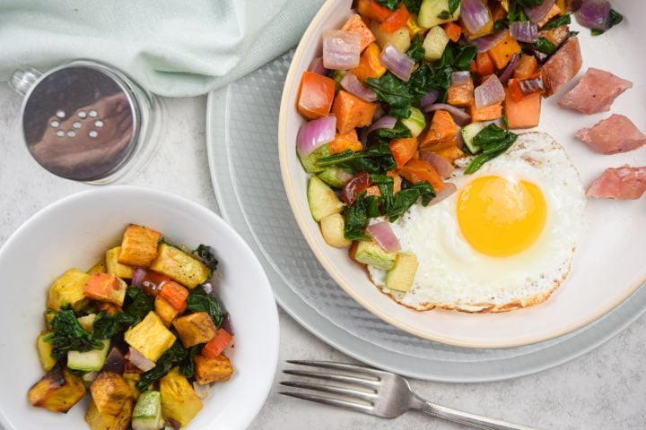 Sweet potato hash with sweet potatoes, zucchini, onions, peppers, and a fried egg on a plate with sliced sausage.