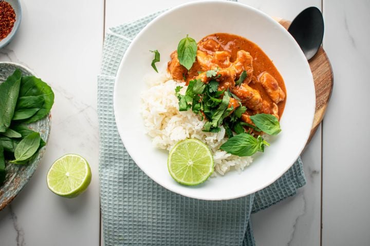 Thai red coconut curry chicken in a bowl with steamed rice, cilantro, basil, and fresh lime.