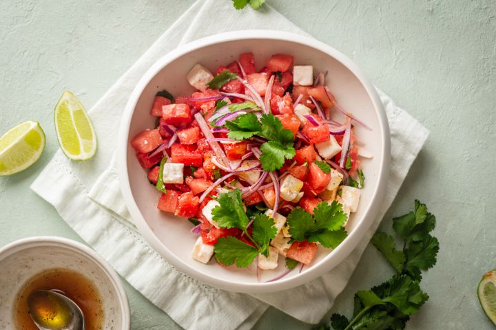 Watermelon and feta salad with cilantro and served with lime vinaigrette in a white bowl with fresh limes on the side.