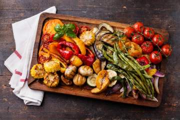 Grilled vegetables including peppers, tomatoes, asparagus, mushrooms, and corn on a wooden board.