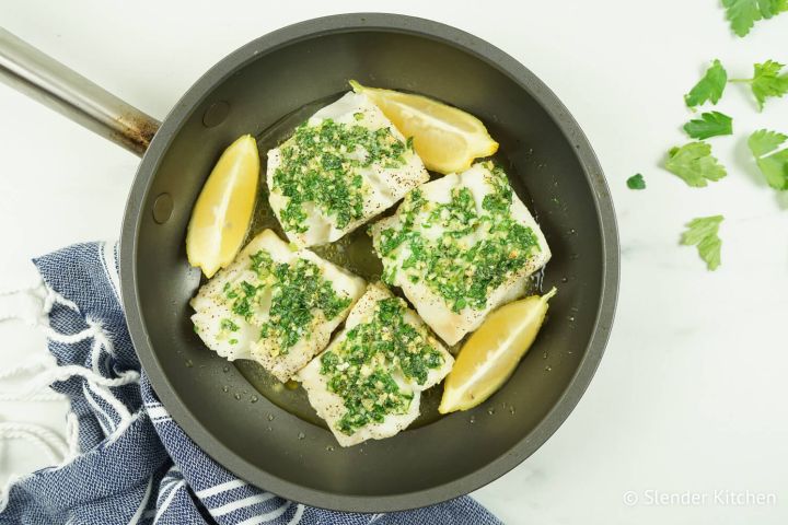 Baked cod with garlic, butter, lemon, and parsley with fresh lemon wedges in a small pan.