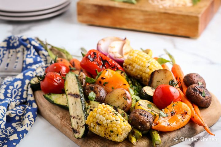 Grilled vegetables including zucchini, peppers, tomatoes, potatoes, and carrots on a wooden cutting board.
