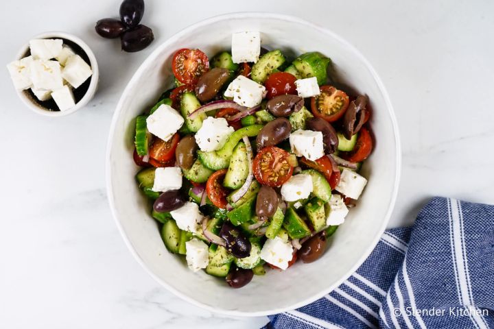 Greek salad with olives, feta cheese, cucumbers, tomatoes, and red onion in a white bowl with a blue napkin. 
