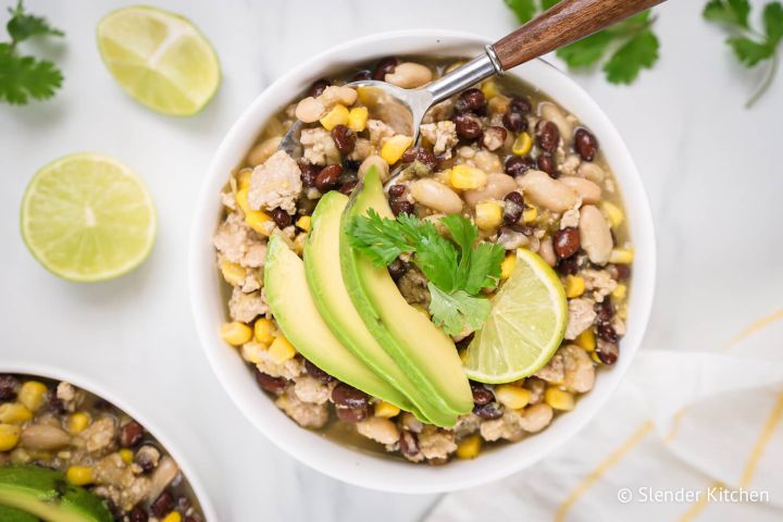 Green turkey chili with ground turkey, black beans, white beans, corn, and sliced avocado in a bowl.