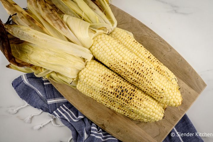 Grilled corn with the husks attached piled on a wood cutting board.