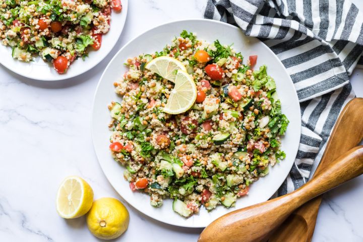 Quinoa tabbouleh with lentils, cucumbers, tomatoes, and parsley served on two plates with lemon.
