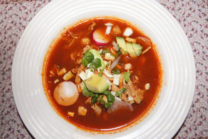 Red chicken pozole in a bowl with shredded chicken, hominy, red chile borth, avocado, and limes. 