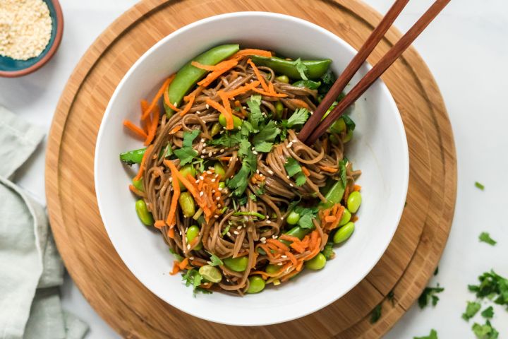 Sesame soba noodles with cilantro, sesame dressing, carrots, edamame, and snap peas in a white bowl with chopsticks.