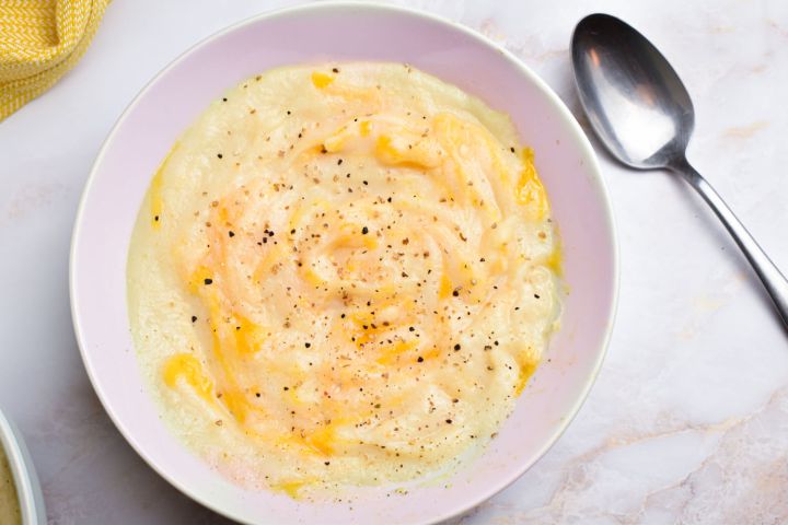 Slow cooker cheesy cauliflower soup with a metal spoon, shredded cheese, and black pepper.