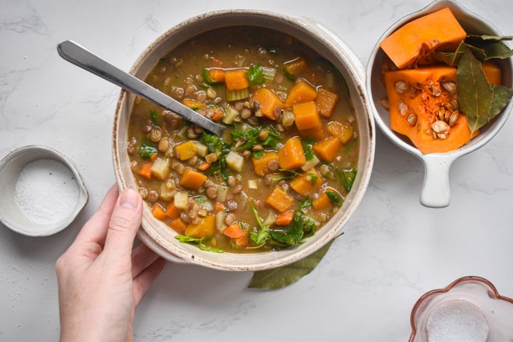 Slow cooker lentil soup with lentils, squash, carrots, spinach, and potatoes in a bowl with a hand holding it.