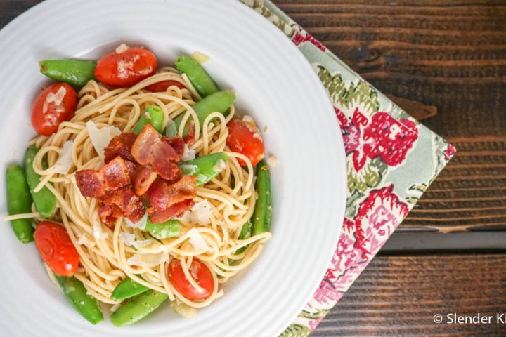 Snap pea pasta with bacon, cherry tomatoes, Parmesan cheese, and lemon in a white bowl.