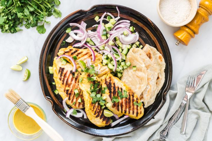Grilled tandoori chicken on a plate with Naan bread, cucumbers, red onions, and cucumbers.