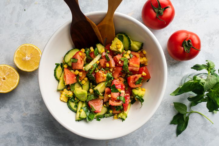 Tomato cucumber salad with corn, avocado, and fresh basil in a bowl with wooden salad tongs.