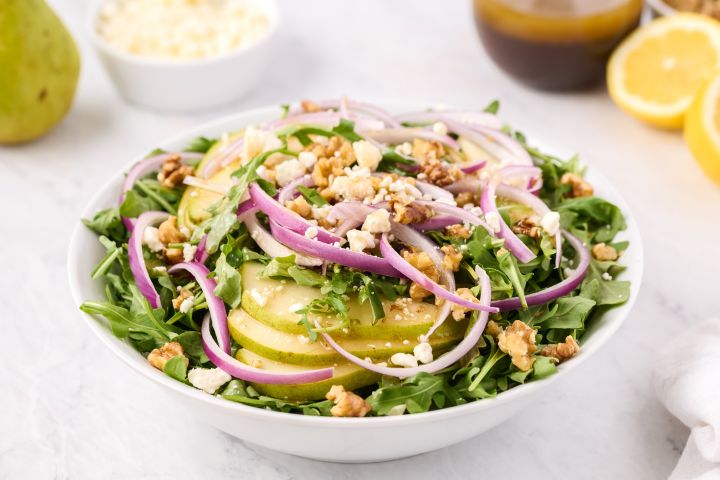 Arugula salad with sliced pears, goat cheese, and red onion tossed with vinaigrette in a white bowl.