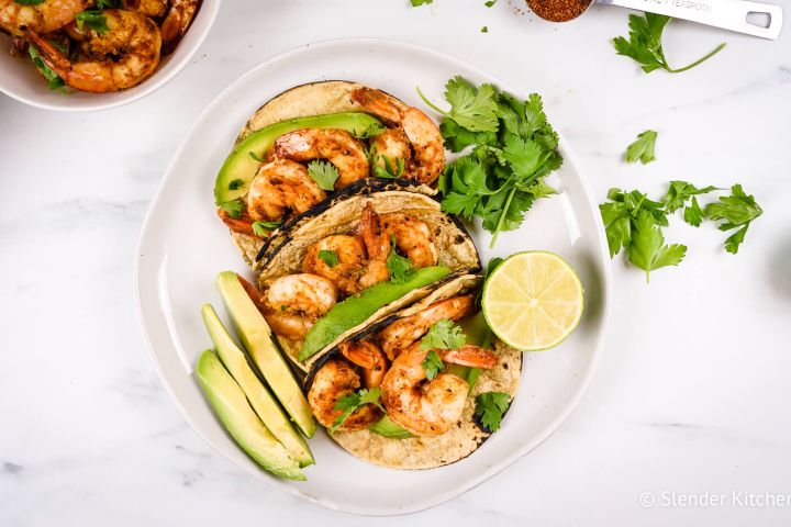 Cajun shrimp tacos in corn tortillas with sliced avocado, fresh lime, and cilantro on a plate.