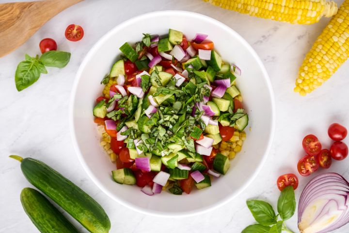 Fresh corn, cucumbers, tomatoes, red onion, and basil in a bowl for corn salad.