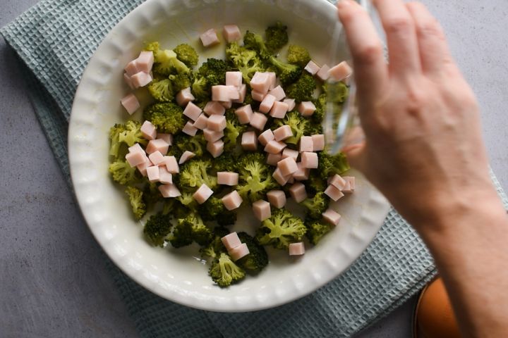 broccoli, ham, and cheese being spread on the bottom of a baking dish.