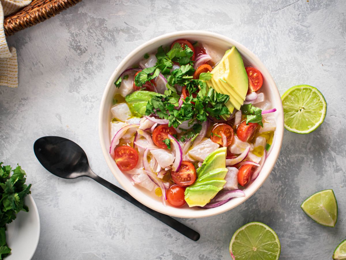 Ceviche with fish marinated in lime juice and served with cilantro, avocado, tomatoes, and red onion in a white bowl.