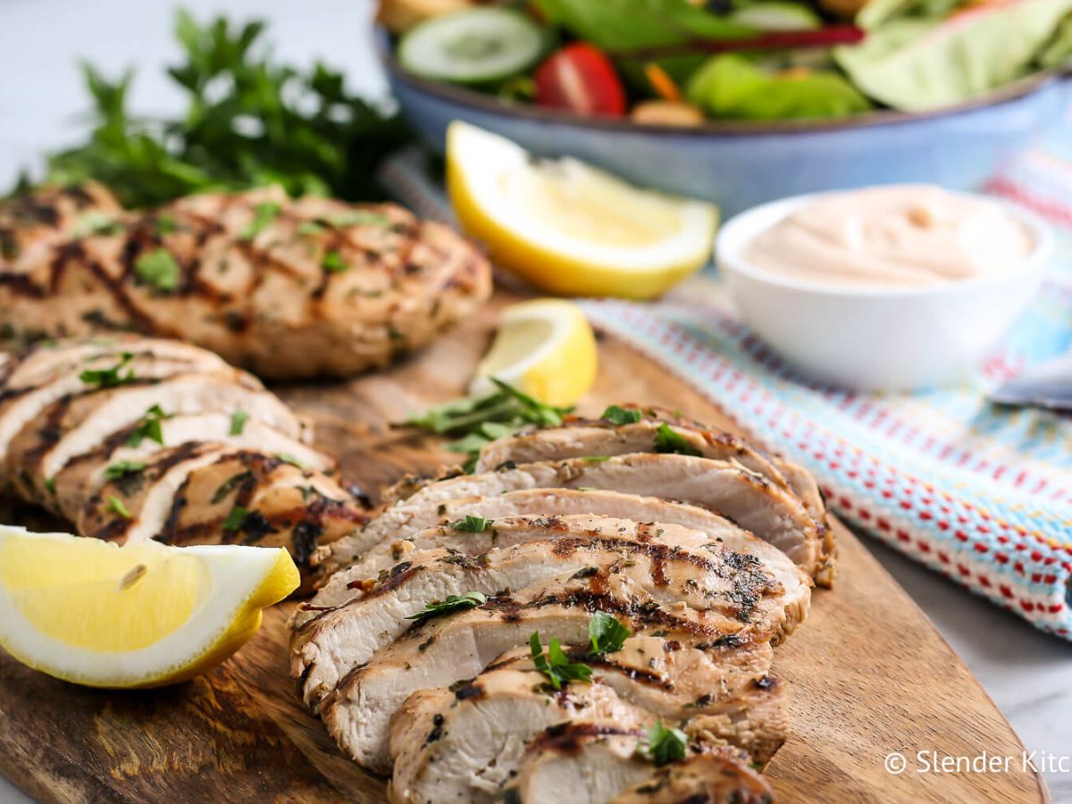 Grilled chicken breast sliced on a wooden cutting board garnished with herbs and lemon