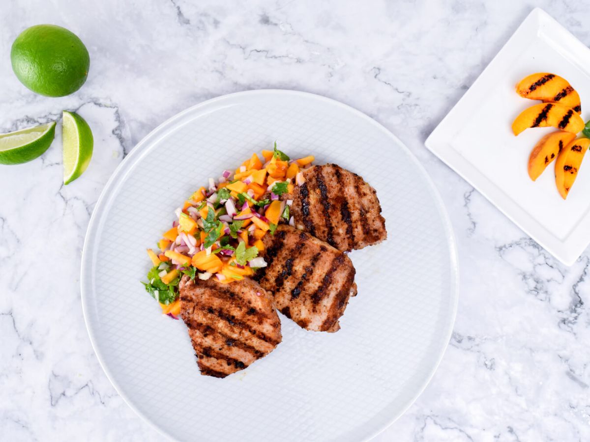 White plate with grilled pork chops accompanied with peach salsa, fresh limes and peaches on the side.