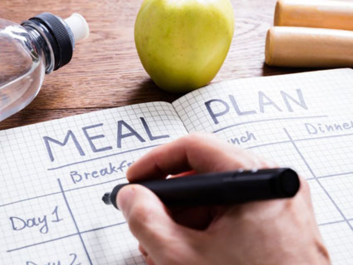 Person writing out a meal plan in a notebook with meals for each day of the week.
