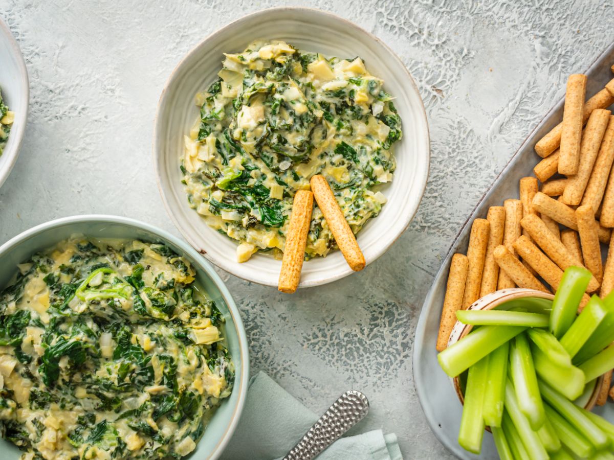 Slow cooker spinach artichoke dip with kale in three bowls with celery and breadsticks.