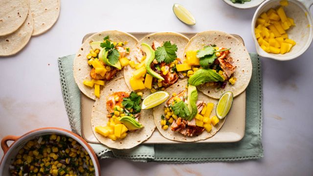 Salmon tacos with corn salsa, mango, fresh avocado slices, and lime wedges on a tray. 